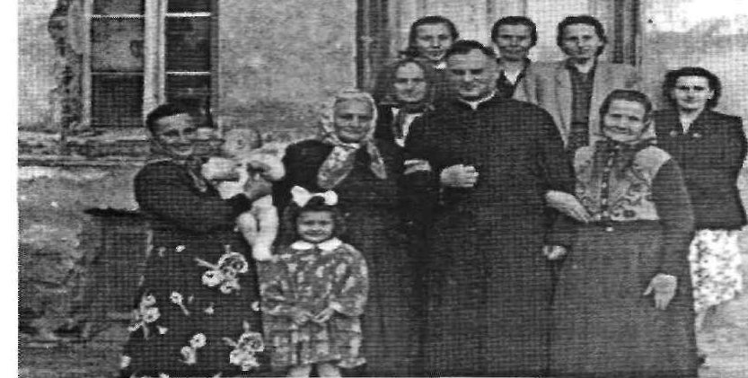 Stare zdjęcie: ksiądz w sutannie stoi w grupie kobiet i dzieci przed murowanym domem.